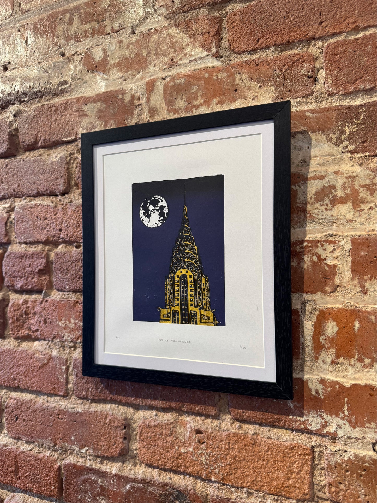 Framed artwork of a building under a moonlit sky on a brick wall.