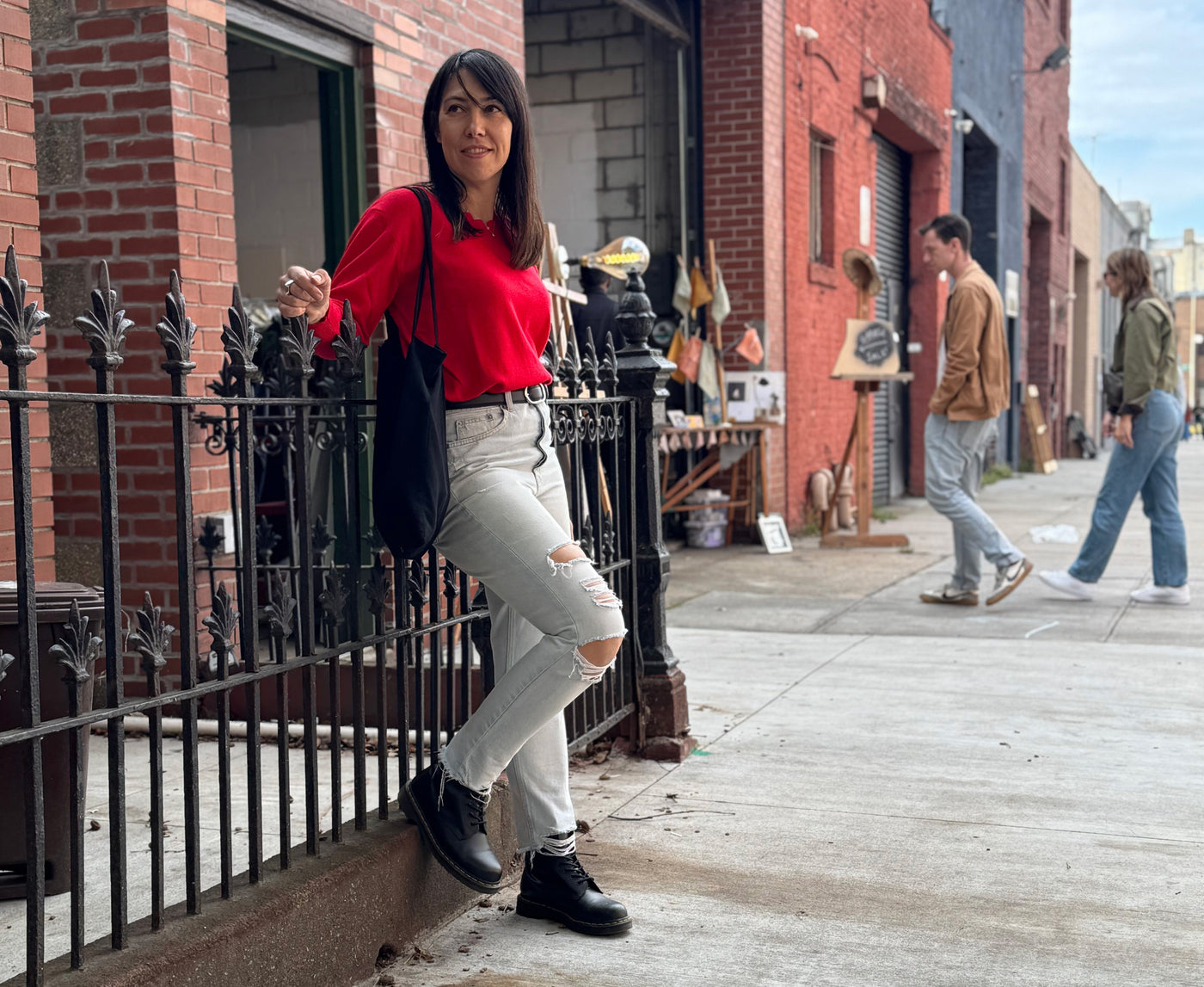 Woman in red shirt and white pants standing on a sidewalk with brick buildings and people in the background.